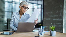 A woman reads a paper document while working in front of her laptop.
